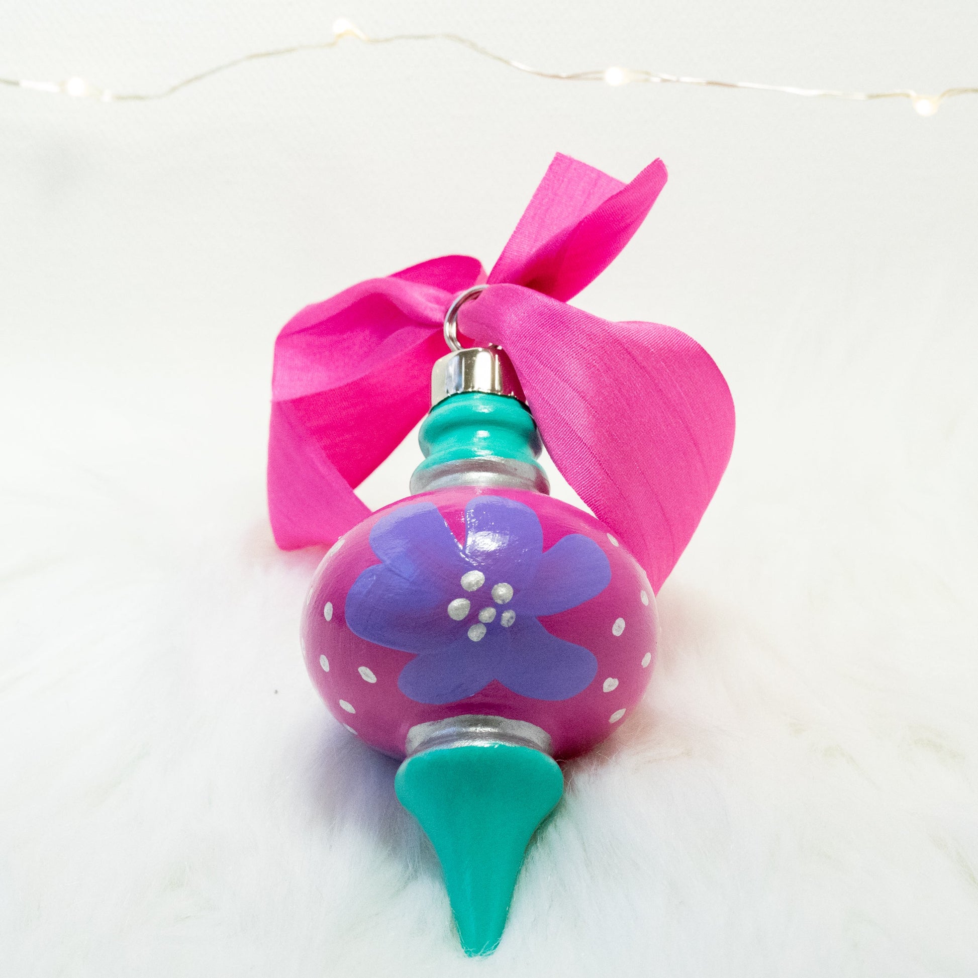 The Maxie Hand Painted Ornament features a fuchsia and bright aqua green base coat, lilac purple flowers with silver accents. Painted using fluid acrylic and acryla gouache paints. Displayed on white faux fur with fairy lights in the background.