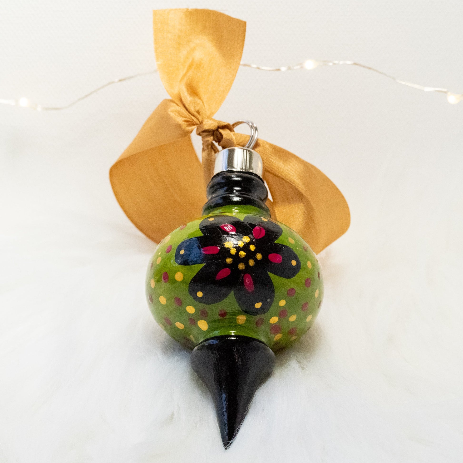 The Sam Hand Painted Ornament features a green gold and black base coat, black flowers with rose violet and gold accents and rose violet and gold polka dots. Painted using fluid acrylic and acryla gouache paints. Displayed on white faux fur with fairy lights in the background.