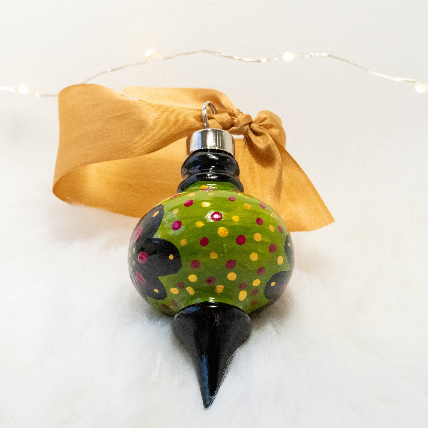 The Sam Hand Painted Ornament features a green gold and black base coat, black flowers with rose violet and gold accents and rose violet and gold polka dots. Painted using fluid acrylic and acryla gouache paints. Displayed on white faux fur with fairy lights in the background.