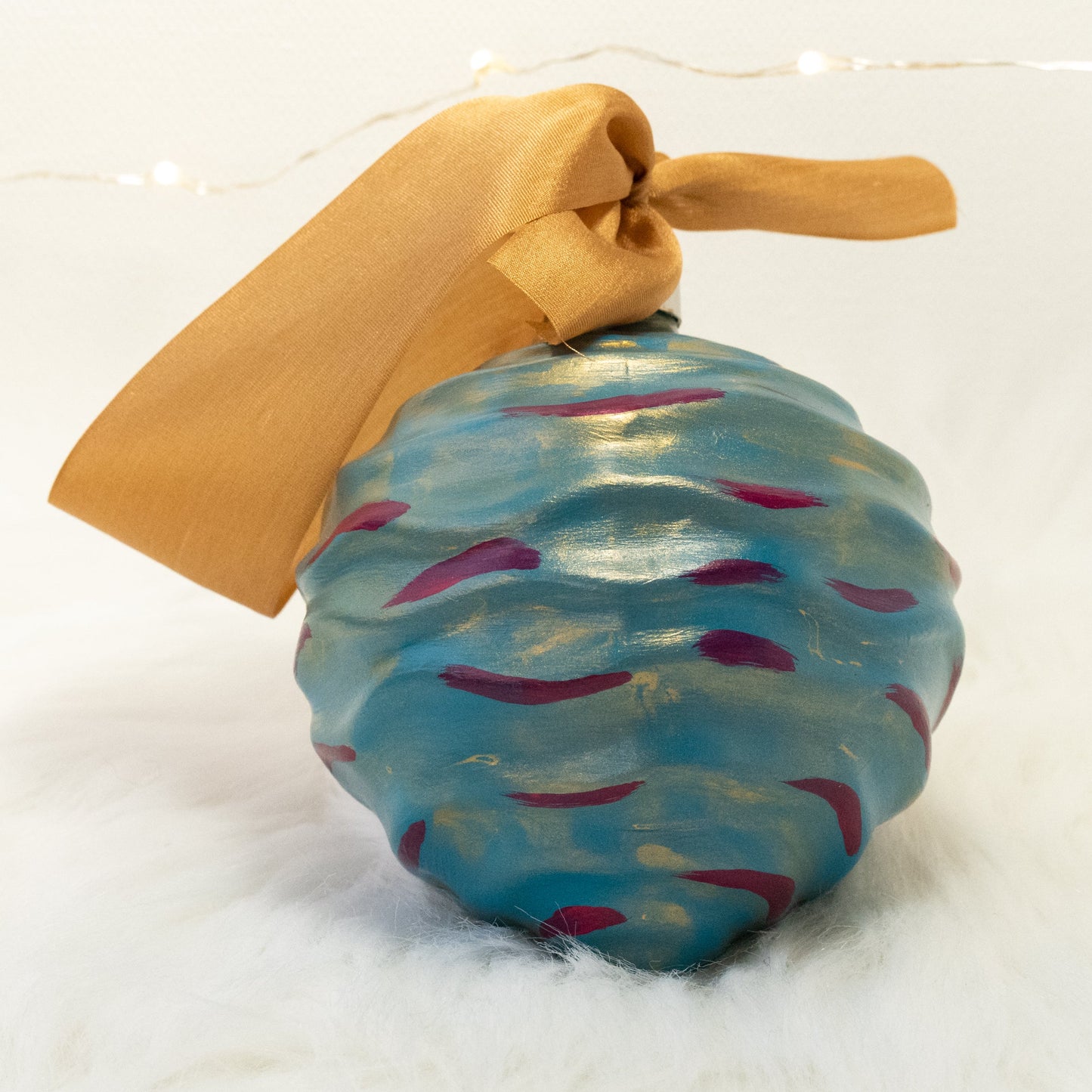 The Stella Hand Painted Ornament features a turquoise blue base coat beneath a transparent gold layer with deep magenta and gold accents. Painted using fluid acrylic and acrylic paints. Displayed on white faux fur with fairy lights in the background.
