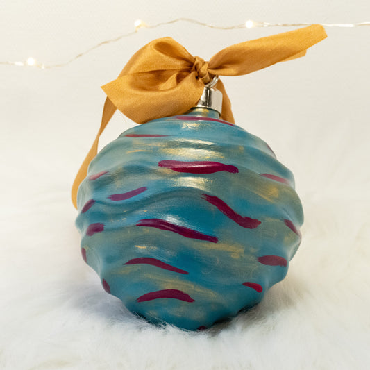 The Stella Hand Painted Ornament features a turquoise blue base coat beneath a transparent gold layer with deep magenta and gold accents. Painted using fluid acrylic and acrylic paints. Displayed on white faux fur with fairy lights in the background.