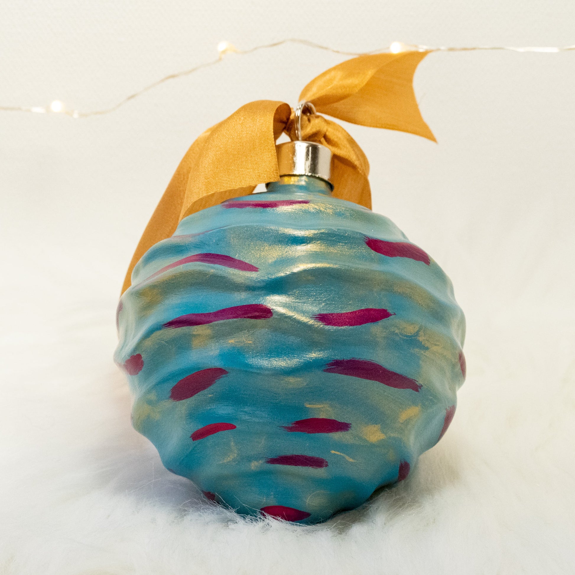 The Stella Hand Painted Ornament features a turquoise blue base coat beneath a transparent gold layer with deep magenta and gold accents. Painted using fluid acrylic and acrylic paints. Displayed on white faux fur with fairy lights in the background.