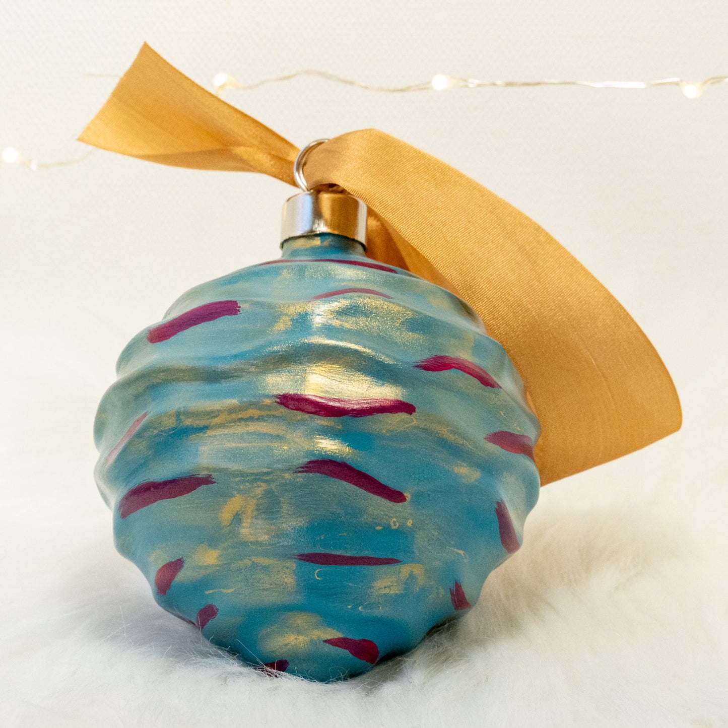 The Stella Hand Painted Ornament features a turquoise blue base coat beneath a transparent gold layer with deep magenta and gold accents. Painted using fluid acrylic and acrylic paints. Displayed on white faux fur with fairy lights in the background.