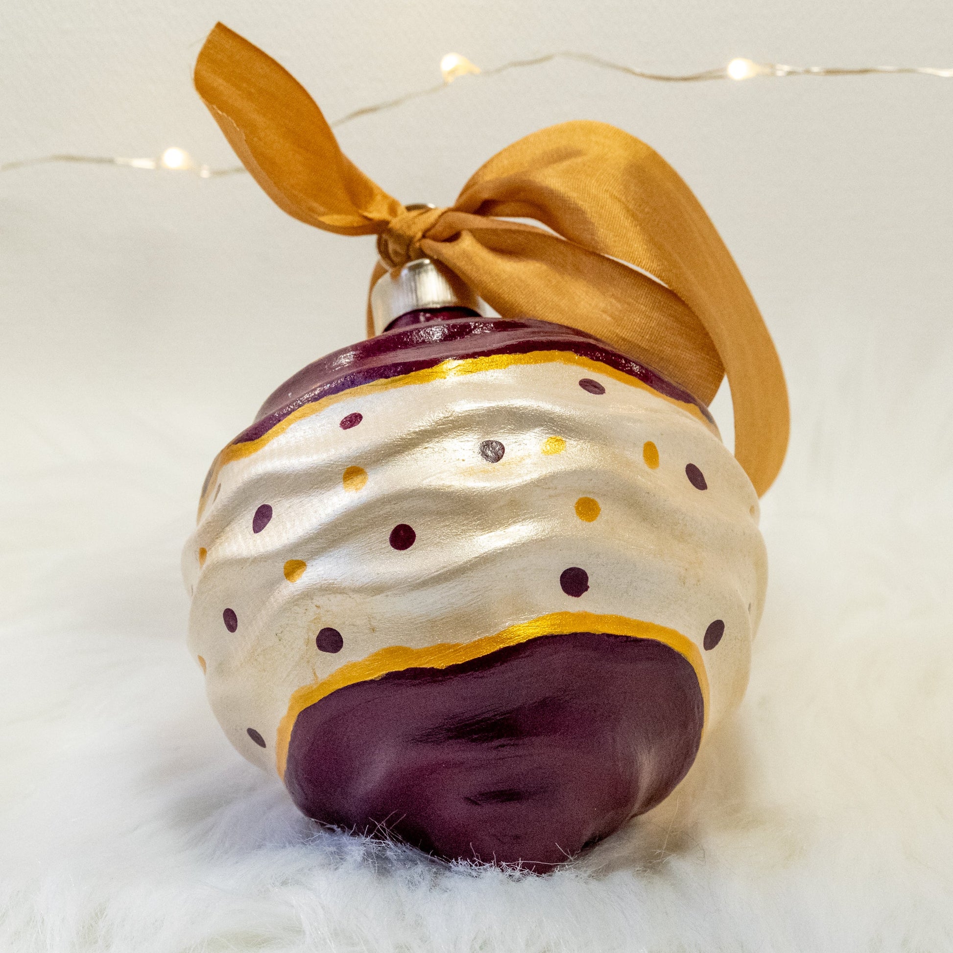 The Tracy Hand Painted Ornament features a white metallic and deep magenta base coat with deep magenta and gold polka dots and gold accents. Painted using fluid acrylic and acryla gouache paints. Displayed on white faux fur with fairy lights in the background.