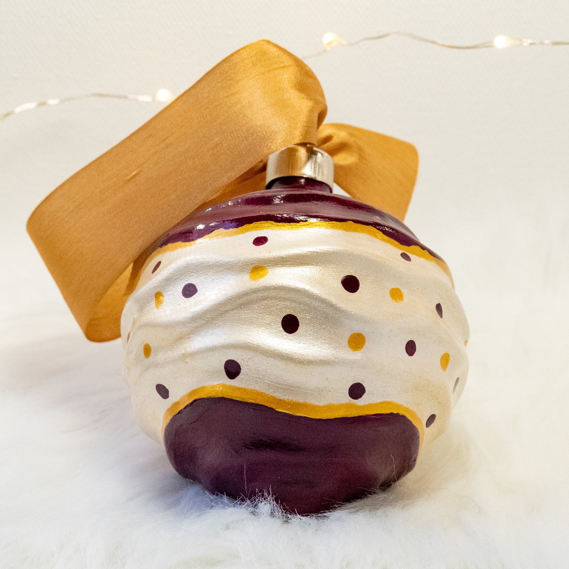 The Tracy Hand Painted Ornament features a white metallic and deep magenta base coat with deep magenta and gold polka dots and gold accents. Painted using fluid acrylic and acryla gouache paints. Displayed on white faux fur with fairy lights in the background.