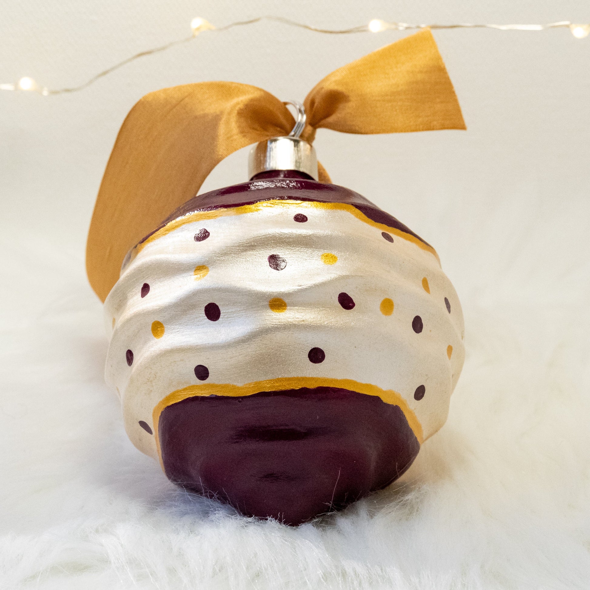 The Tracy Hand Painted Ornament features a white metallic and deep magenta base coat with deep magenta and gold polka dots and gold accents. Painted using fluid acrylic and acryla gouache paints. Displayed on white faux fur with fairy lights in the background.