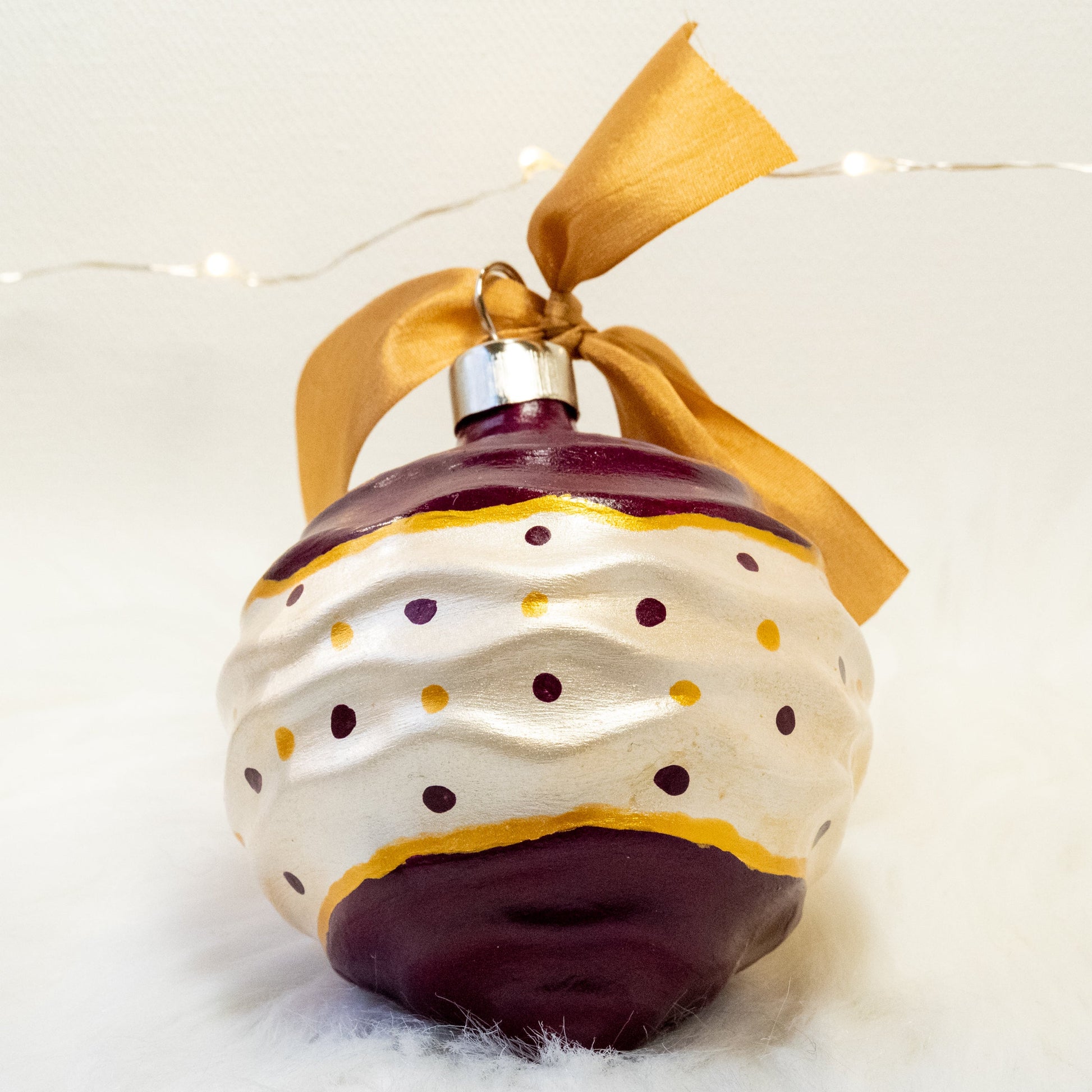 The Tracy Hand Painted Ornament features a white metallic and deep magenta base coat with deep magenta and gold polka dots and gold accents. Painted using fluid acrylic and acryla gouache paints. Displayed on white faux fur with fairy lights in the background.