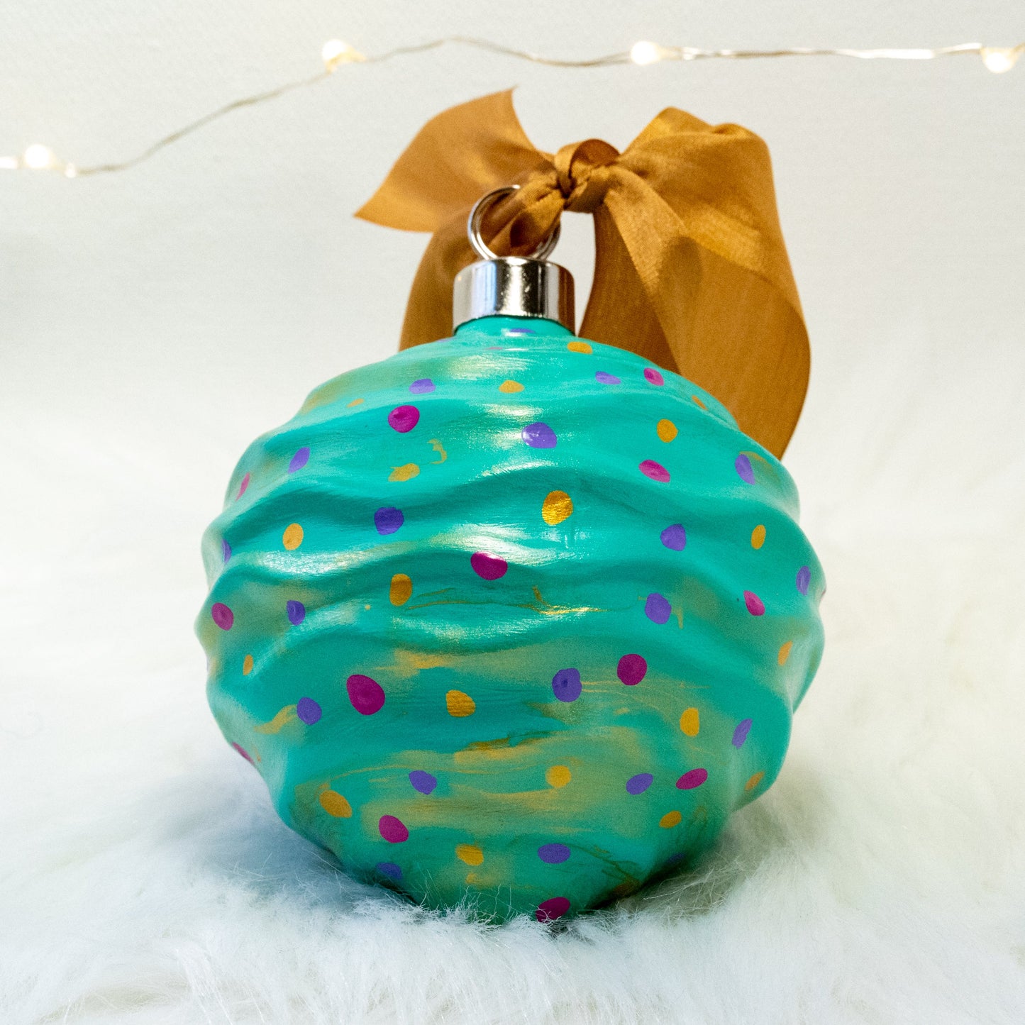 The Trina Hand Painted Ornament features a bright aqua green base coat with fuchsia, purple and gold polka dots and gold accents. Painted using fluid acrylic and acrylic paints. Displayed on white faux fur with fairy lights in the background.