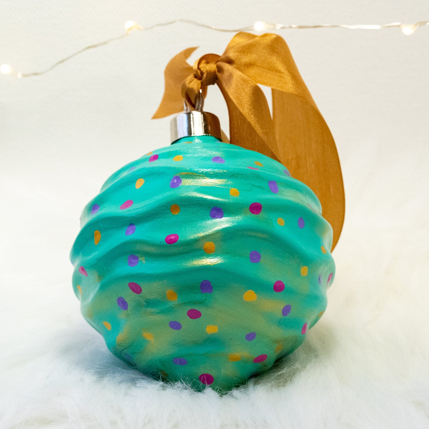 The Trina Hand Painted Ornament features a bright aqua green base coat with fuchsia, purple and gold polka dots and gold accents. Painted using fluid acrylic and acrylic paints. Displayed on white faux fur with fairy lights in the background.