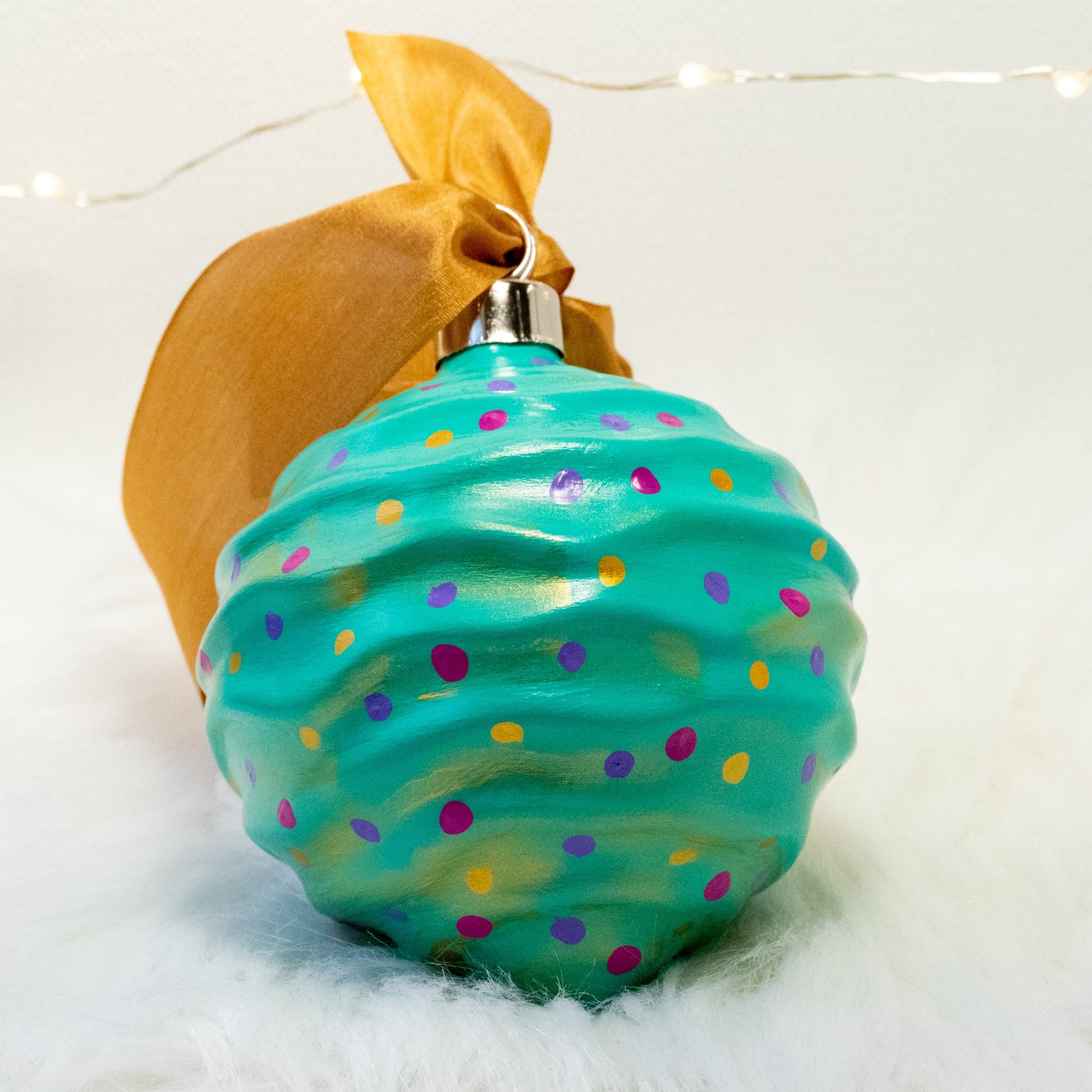 The Trina Hand Painted Ornament features a bright aqua green base coat with fuchsia, purple and gold polka dots and gold accents. Painted using fluid acrylic and acrylic paints. Displayed on white faux fur with fairy lights in the background.